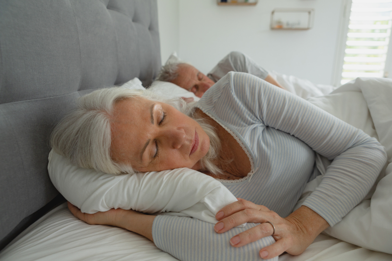 3 Health Benefits of Using Your AC While Sleeping. Photo of a an elderly couple sleeping soundly in their bed.